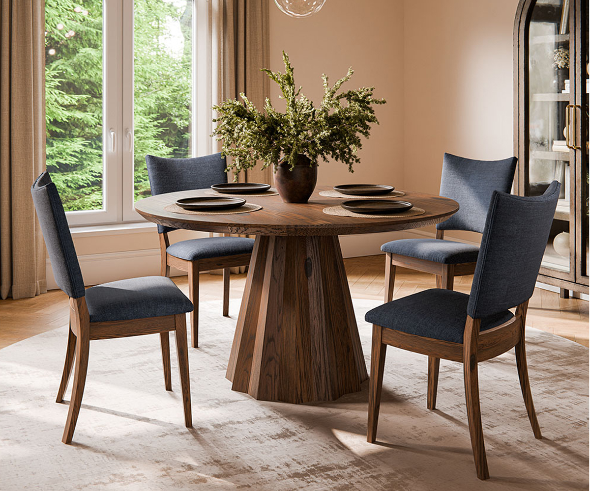 A round wooden pedestal table surrounded by four blue-upholstered chairs, with a vase of greenery on the table, set in a light-filled dining room near a large window.